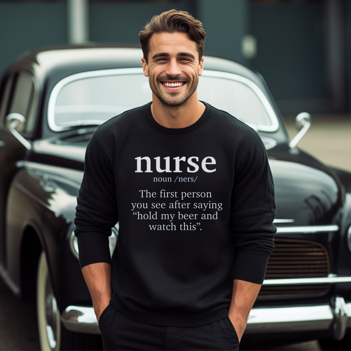 A man wearing a black sweatshirt with a funny nurse-themed shirt design, reading: 'The first person you see after saying “hold my beer and watch this”.' He is standing in front of a classic car, smiling.