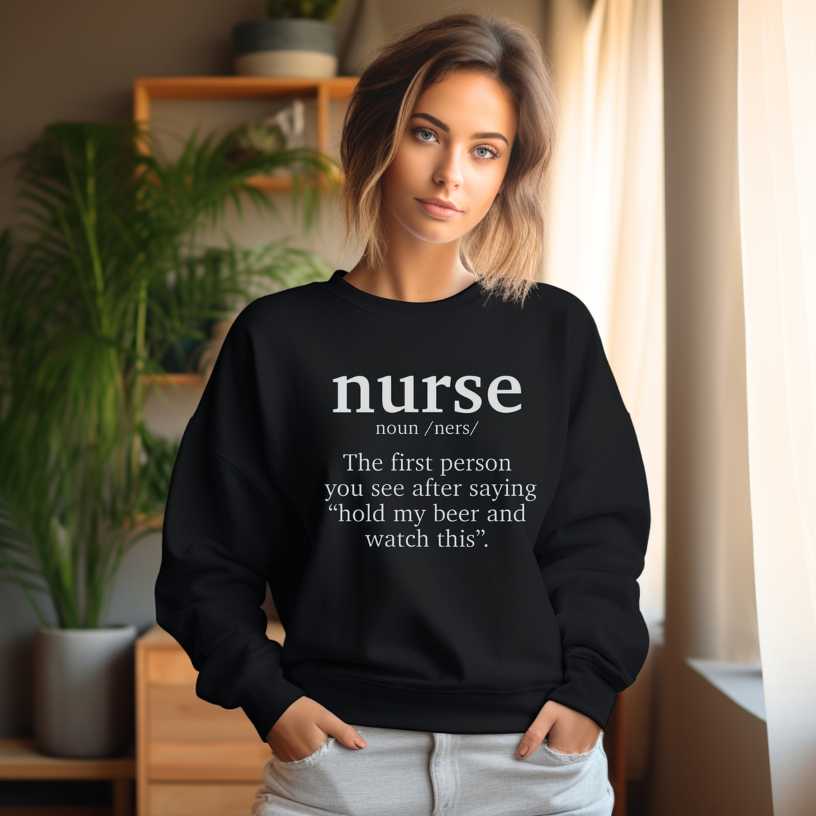 A woman wearing a black sweatshirt with a funny nurse-themed shirt design, reading: 'The first person you see after saying “hold my beer and watch this”.' She is standing in a well-lit room with plants and shelves in the background.