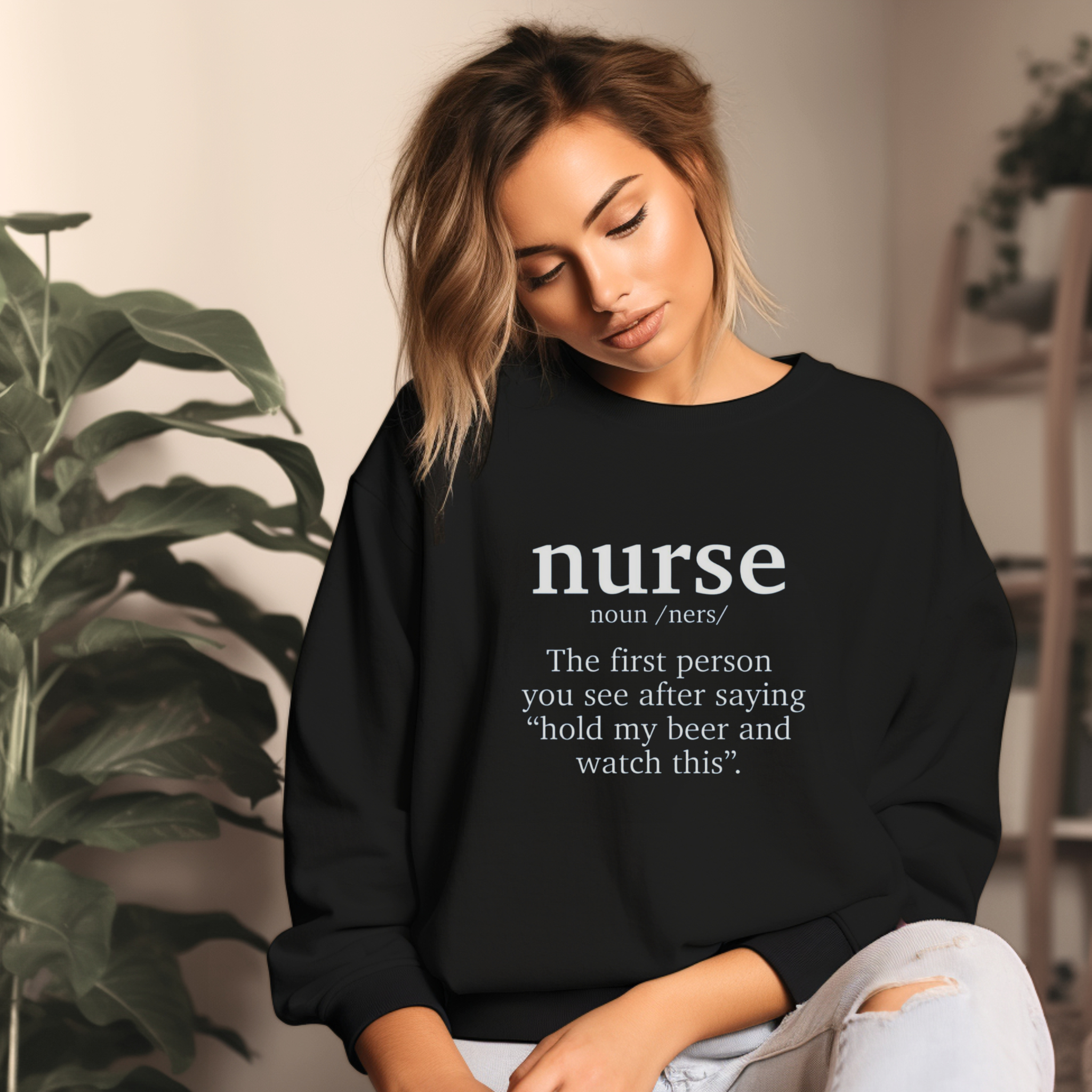 A woman wearing a black sweatshirt with a funny nurse-themed shirt design, reading: 'The first person you see after saying “hold my beer and watch this”.' She is sitting in a cozy room with plants in the background.