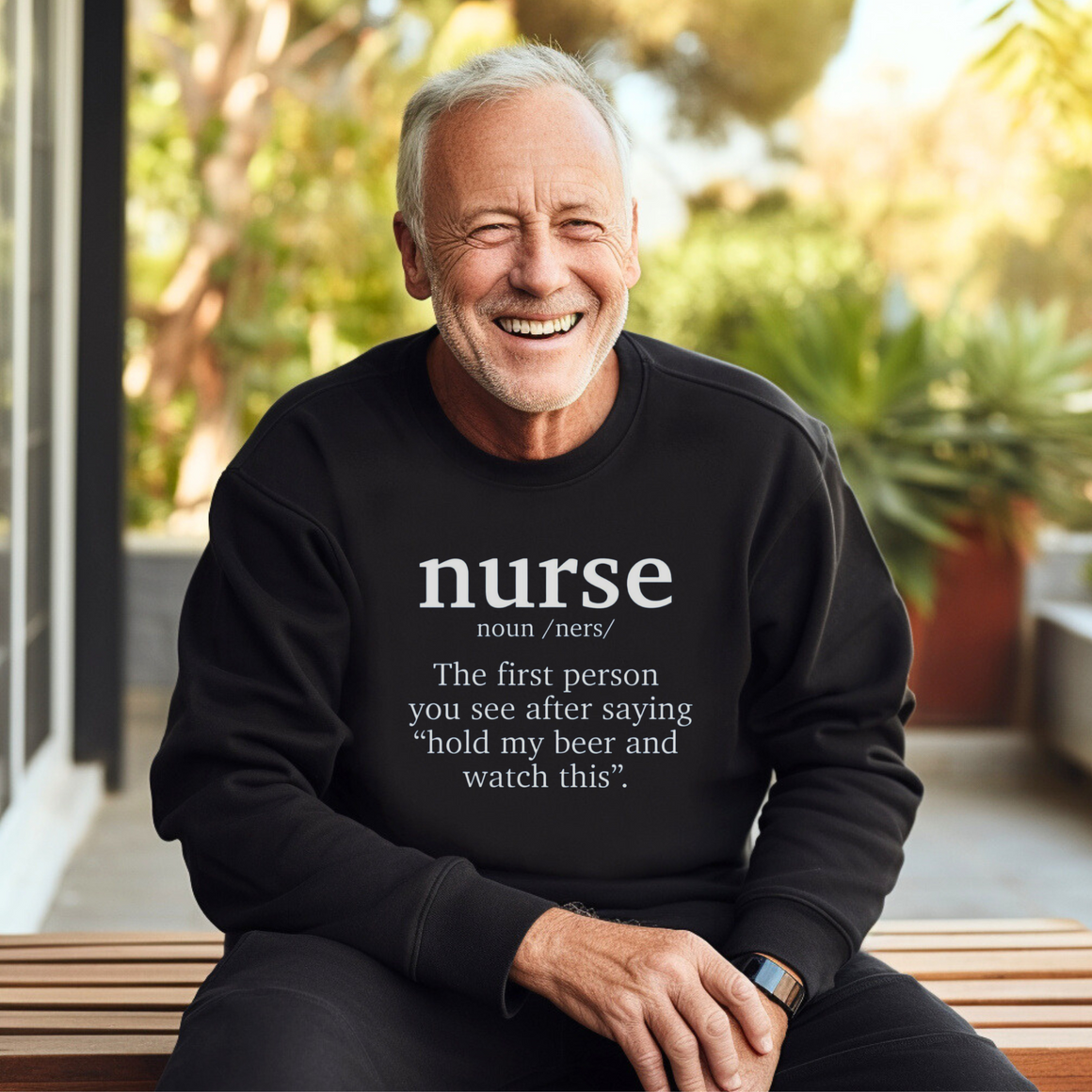 An older man wearing a black sweatshirt with a funny nurse-themed shirt design, reading: 'The first person you see after saying “hold my beer and watch this”.' He is sitting outdoors, smiling, with greenery in the background