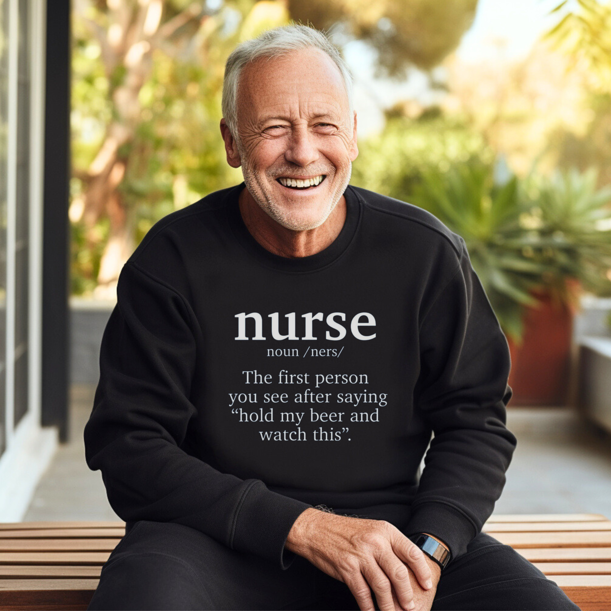 An older man wearing a black sweatshirt with a funny nurse-themed shirt design, reading: 'The first person you see after saying “hold my beer and watch this”.' He is sitting outdoors, smiling, with greenery in the background