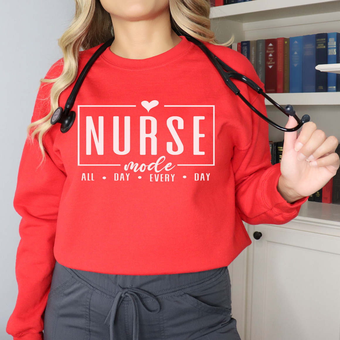 Woman wearing a red sweatshirt with 'Nurse Mode All Day Every Day' printed in white text, a stethoscope around her neck, and hands on her waistband. Stylish design perfect for Nurses Week Custom Shirt Suggestions.