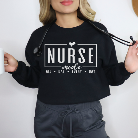 Woman wearing a black sweatshirt with 'Nurse Mode All Day Every Day' printed in white text. She holds a coffee mug in one hand and a stethoscope draped around her neck. Stylish design ideal for Nurses Week Custom Shirt Suggestions.