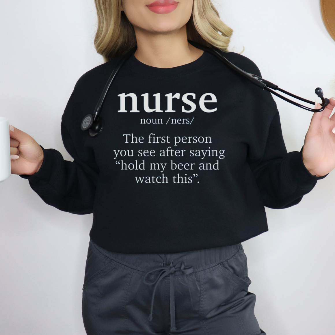 Woman wearing a black sweatshirt with the humorous text 'nurse: The first person you see after saying hold my beer and watch this.' Ideal for those looking for funny nurse-themed shirts that add a touch of humor to their wardrobe.