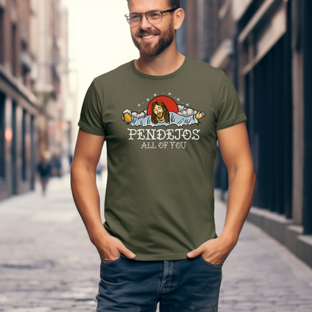 Man standing on a city street wearing an olive green t-shirt with a humorous graphic of a figure resembling Jesus holding a beer mug, surrounded by clouds and stars. The text on the shirt reads 'Pendejos All of You.' The man is smiling, with his hands in his pockets.