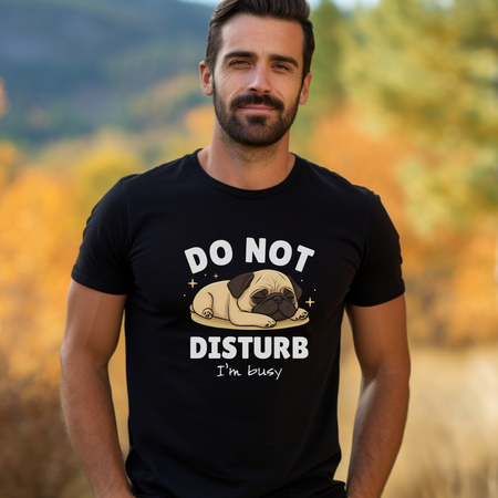 A man wearing a black t-shirt with a cute sleeping pug illustration and the text "DO NOT DISTURB, I'm busy" printed on it. He is standing outdoors with a backdrop of autumn foliage and mountains.