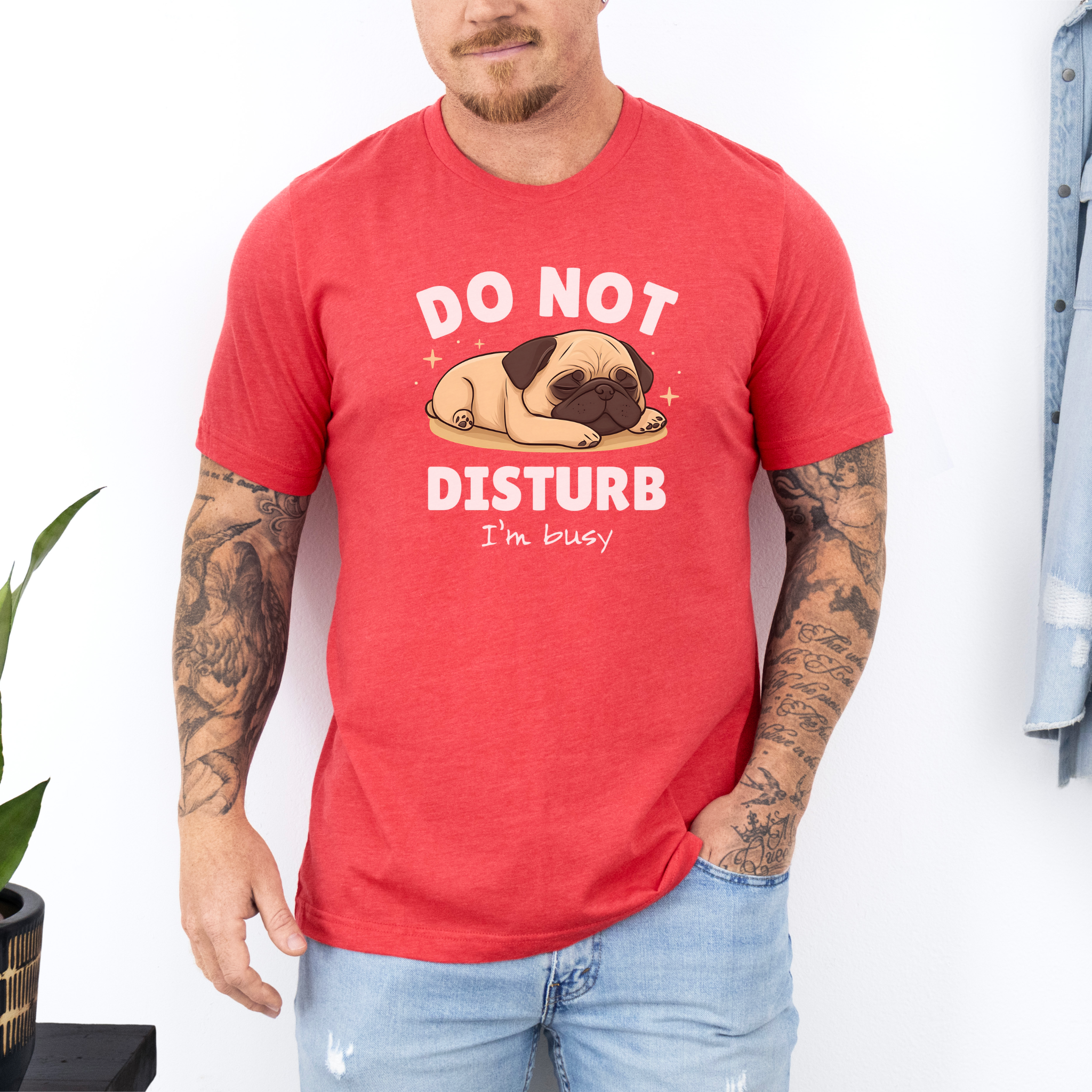 A man wearing a heather red t-shirt with a cute sleeping pug illustration and the text "DO NOT DISTURB, I'm busy" printed on it. He has tattoos on his arms and is standing indoors with a hand in his pocket.