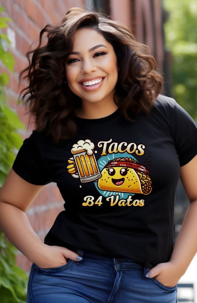 A smiling woman with curly dark brown hair wears a black T-shirt that features a cheerful, cartoon-style taco character wearing a red bandana and a foamy beer mug with a handle. The T-shirt text reads "Tacos B4 Vatos" in bold, playful font, with the taco and beer images adding a fun, lighthearted vibe. The woman stands against a brick wall with greenery in the background, looking relaxed and happy.