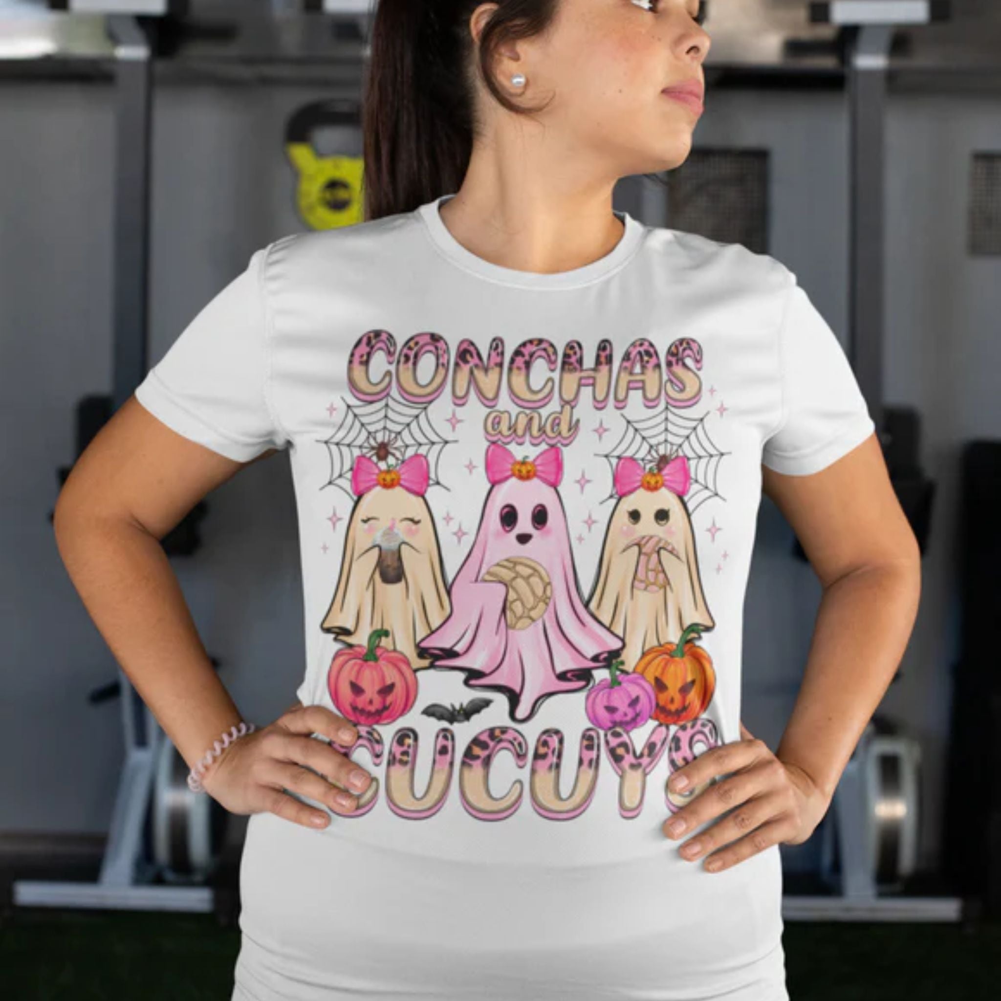 Woman wearing a white t-shirt featuring the "Conchas and Cucuy's" design, part of our Latino-inspired Halloween apparel. The shirt displays three cute ghost figures holding conchas and decorated with pink bows, spider webs, and festive pumpkin accents, combining Halloween spirit with Día de los Muertos charm. This unisex, soft-style shirt offers a unique blend of spooky and sweet for the Halloween season.